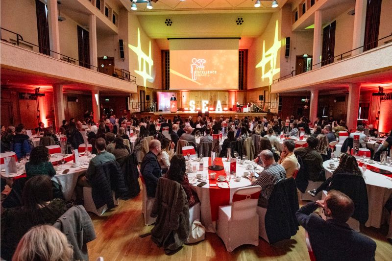 Wide view of the Staff Excellence Awards ceremony in a large hall, with guests seated at round tables watching the stage where the “SEA” letters and event screen are illuminated.